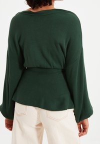 Dunkelgrüner, strukturierter Strickpullover mit lockerem Schnitt und ausgestellten Ärmeln, in der Taille gerafft. Kombiniert mit hellen, hoch taillierten Hosen.