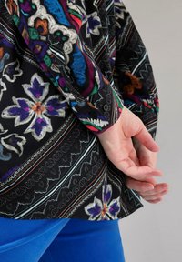 Camicia nera a fantasia con design floreali colorati, con polsini abbottonati e orlo dritto. Indossata con pantaloni blu, mani incrociate dietro.