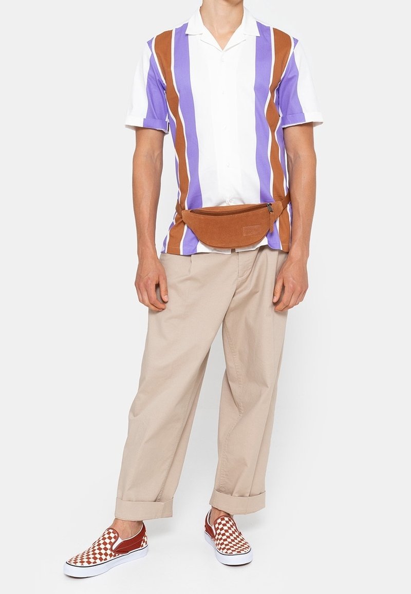 Homme portant une chemise à manches courtes à rayures verticales, un pantalon beige, un sac banane marron et des chaussures slip-on à carreaux rouges et blancs.
