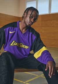 Violeta un melna Nike krekls ar "LAKESHOW" logo; pievienoti dzelteni akcenti. Valkāts ar melnām biksēm, kurām ir teksturēts ziedu raksts.