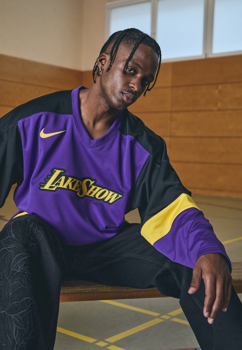 Violeta un melna Nike krekls ar "LAKESHOW" logo; pievienoti dzelteni akcenti. Valkāts ar melnām biksēm, kurām ir teksturēts ziedu raksts.