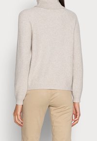 Maglione beige lavorato a maglia con collo alto, texture a coste e maniche aderenti, abbinato a pantaloni marrone chiaro, visto da dietro.