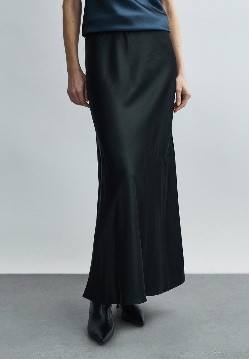 Kobieta ubrana w długą, czarną jedwabną spódnicę, zestawioną z czarnymi, spiczastymi botkami, stoi na szarej podłodze na tle jednolitej ściany.