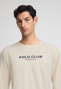 Maglietta a maniche lunghe color crema realizzata in morbido cotone, con ricamo "POLO CLUB" in blu scuro e rosso, colletto rotondo e vestibilità comoda.