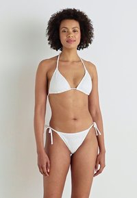 Hvit bikini-sett med en teksturert trekantet topp og lavt liv-bunn med justerbare snorer på sidene. Enkel og minimalistisch design.