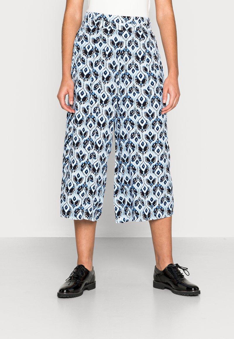 Femme portant un pantalon large bleu et noir à motifs et coupé au niveau des mollets, avec des chaussures noires à lacets, debout devant un fond clair uni.