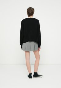 Černý oversized pletený svetr, šedá skládací sukně a černé vysoké tenisky. Jednoduché pozadí a model otočený zády k fotoaparátu.