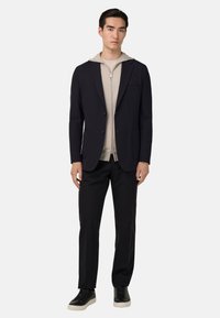 Námorník s přiléhavým střihem, na dva knoflíky a s našitými kapsami, přes béžovou mikinu na zip. Tmavé kalhoty a černé tenisky dotvářejí vzhled.