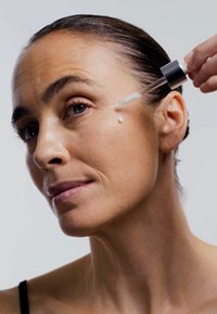 Bottiglia contagocce che distribuisce un siero trasparente sulla pelle vicino all'occhio. Il soggetto ha una pelle liscia e leggermente strutturata e i capelli raccolti.