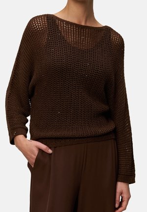 Pull marron foncé en maille ouverte à manches longues porté sur une couche sombre, assorti d’un pantalon marron ample assorti, une main dans la poche.