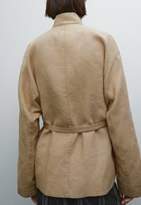 Vaahto hiekka puuvillatakki, jossa on vyöllinen vyötärö, leveät hihat ja korkea kaulus. Takissa on sileä, teksturoitu kangas ja rento istuvuus.