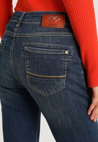 Jeans de denim de color azul oscuro con una cintura alta, que cuentan con una parche de marca de cuero, bolsillos traseros cosidos y detalles en hilo dorado.