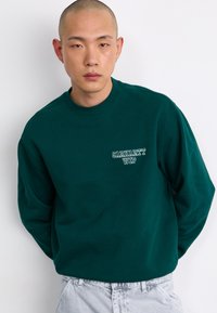 Grøn bomulds sweatshirt med rund hals og lange ærmer, prydet med et lille hvidt trykt logo på venstre bryst. Struktureret stof.