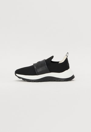 Sort slip-on sneaker med tykk hvid sål, strikket overdel, bred elastisk rem over mellemfoden og trækstrop ved hælen.