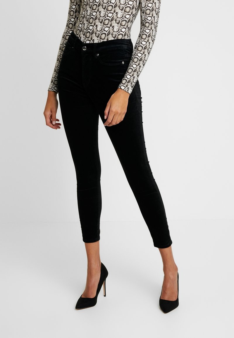 Femme portant un pantalon skinny noir, des talons hauts noirs et un haut à imprimé serpent à manches longues, se tenant devant un fond neutre.