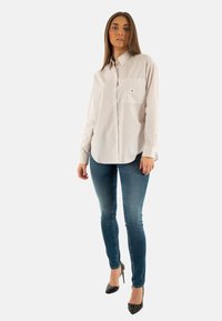 Camisa blanca a rayas con botones y un bolsillo, combinada con unos pantalones ajustados de mezclilla azul y tacones altos negros, mostrando un diseño elegante y moderno.