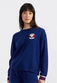 Marineblauwe trui met geribbelde ronde hals en lange mouwen. Kenmerkt een geborduurde Snoopy-afbeelding en een rood-wit cirkelvormig detail op de borst.