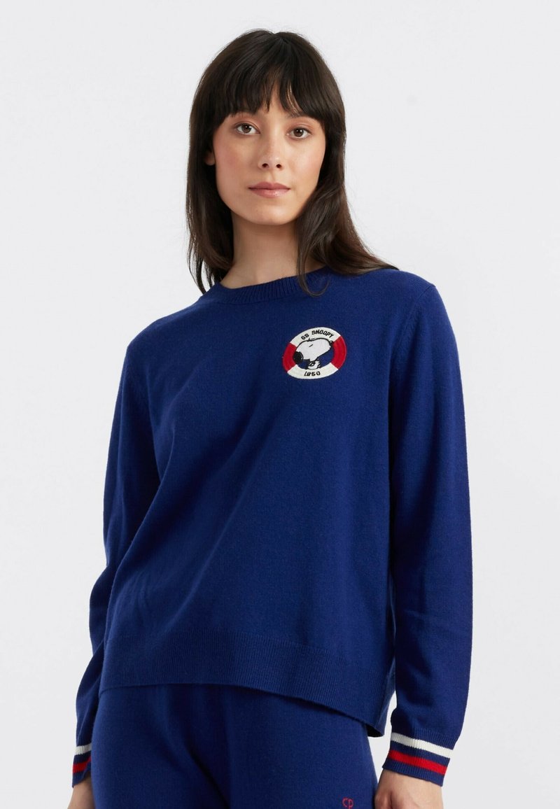 Marineblauwe trui met geribbelde ronde hals en lange mouwen. Kenmerkt een geborduurde Snoopy-afbeelding en een rood-wit cirkelvormig detail op de borst.