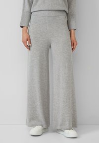 Personne portant un pantalon en tricot large gris clair, un pull assorti et des baskets blanches sur un fond gris uni.