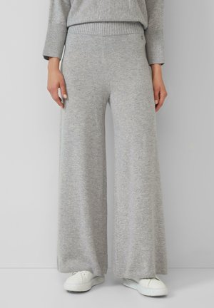 Persona che indossa pantaloni a gamba larga in maglia grigia chiara, un maglione abbinato e sneakers bianche su uno sfondo grigio uniforme.