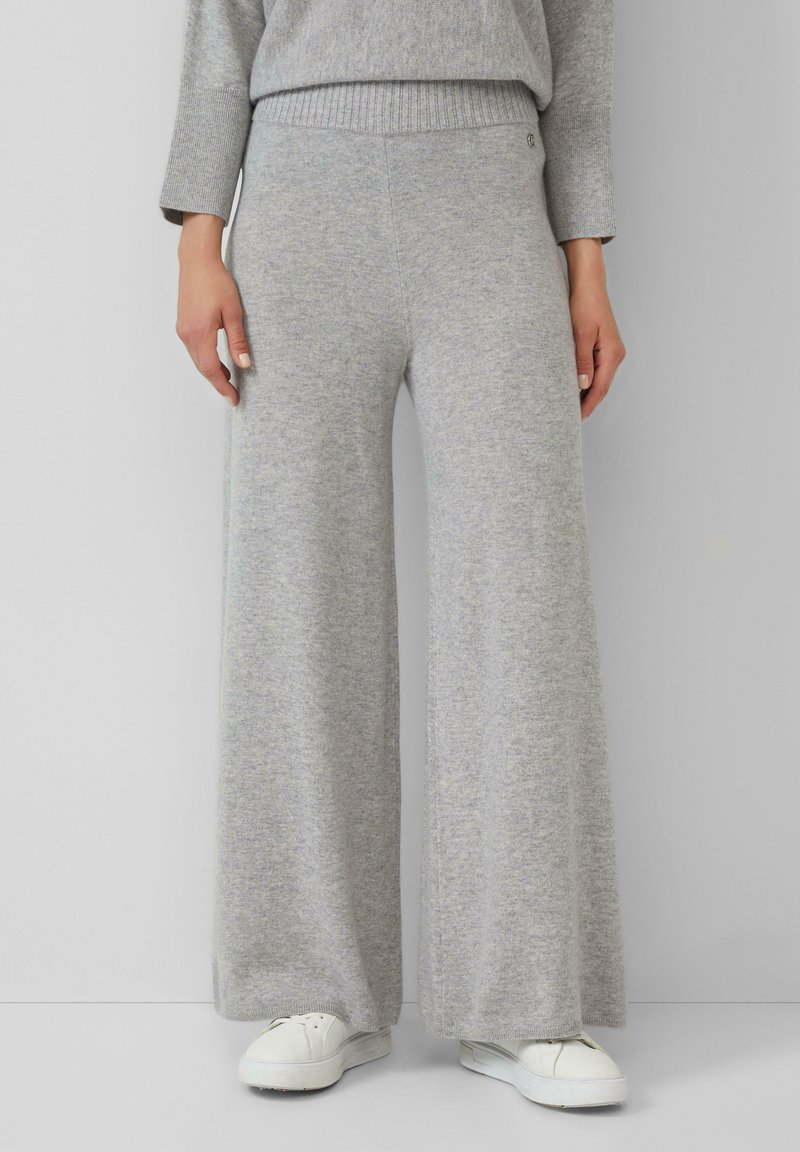 Personne portant un pantalon en tricot large gris clair, un pull assorti et des baskets blanches sur un fond gris uni.
