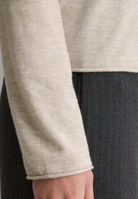 Beżowy sweter z dzianiny, z miękką teksturą i zawiniętą mankietą, w połączeniu z ciemnoszarym materiałem w prążki poniżej.