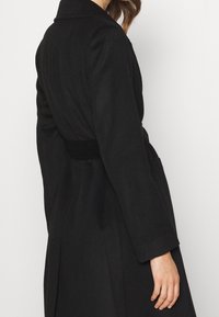 Svart ullkappa med långa ärmar, ett bälte i midjan och en stor krage. Har en slät yta och två sidfickor.