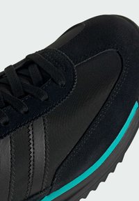 Black athletic shoe featuring a combination of smooth nylon and suede materials, with a teal accent on the midsole and black laces.