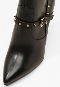 Bottine en cuir noir à bout pointu, avec une sangle ornée de clous dorés et un détail de boucle, texture lisse et design ajusté.