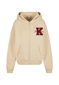 Bež dukserica sa zipom, sa prednjim džepom, izvezenim crvenim slovom "K" i tekstom u kurzivu. Ima kapuljaču sa vezicom i rebraste manžete.