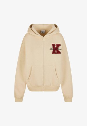 Bežs kapuces džemperis ar rāvējslēdzēju, ar priekšējo kabatu, izšūtu sarkano burtu "K" un rakstītu tekstu kursīvā. Iespējas ietver kapuci ar auklu un ribotu piedurkni.