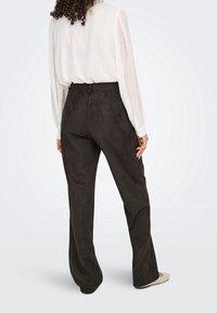 Femme aux cheveux bouclés portant un chemisier blanc transparent à manches longues et un pantalon noir ample taille haute, debout de dos au spectateur.