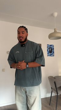 Överdimensionerad grå skjorta med ficka och korta ärmar, kombinerad med ljusblå slitna jeans. Inomhusmiljö med minimal inredning.