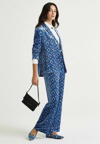 Completo blu a fantasia con motivo a diamante, realizzato in materiale liscio. Abbinato a una camicia bianca, una borsa nera e scarpe blu coordinate.