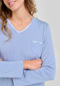 Mujer sonriendo con los brazos cruzados, vestida con una camiseta de manga larga de color azul claro con un escote en V ribeteado en satén y un pequeño bolsillo en el pecho.