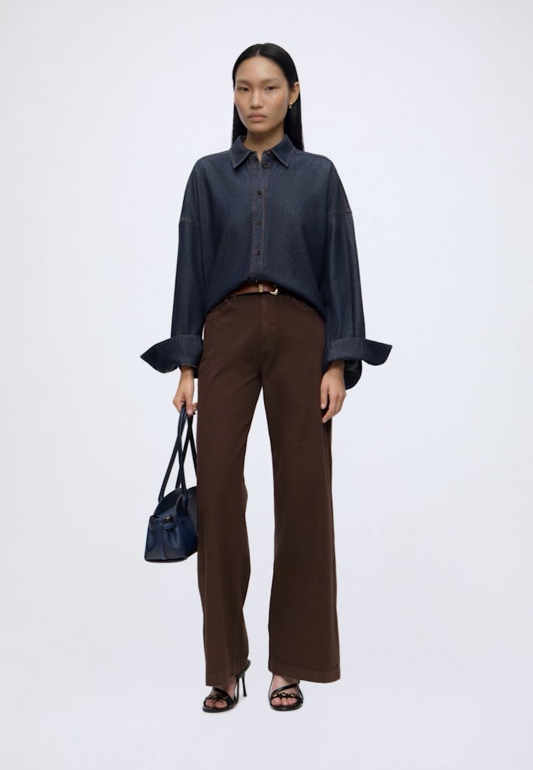 Femme aux cheveux noirs et raides porte une chemise ample en jean foncé, un pantalon marron à jambes larges, des talons noirs, et tient un petit sac à main bleu marine sur fond blanc.