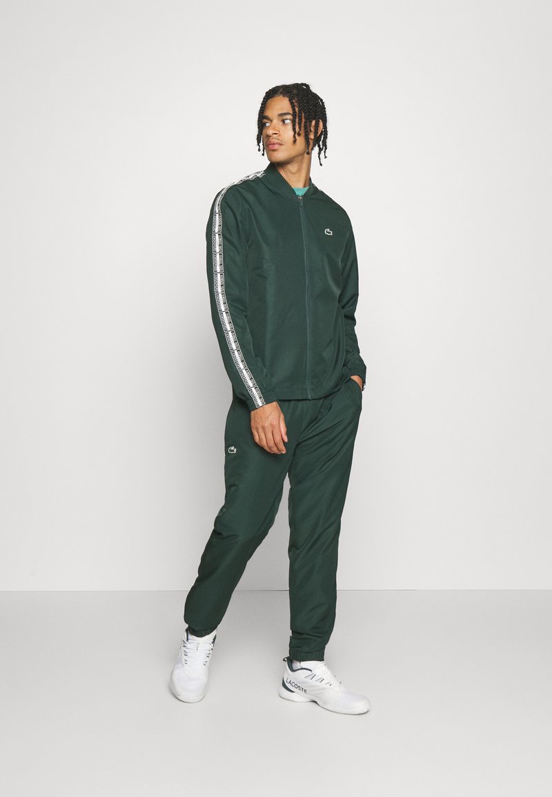 Chándal deportivo verde en tejido suave, que cuenta con una chaqueta con cremallera y franjas laterales blancas, y pantalones ajustados a juego. Los detalles incluyen un pequeño logo.