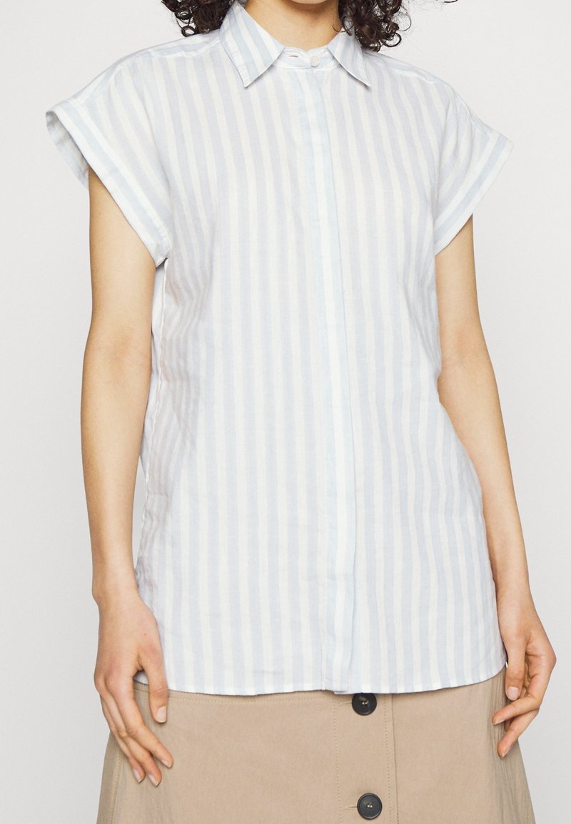 Femme portant une chemise bleu clair et blanche à rayures verticales, manches courtes, avec col, et une jupe beige boutonnée.