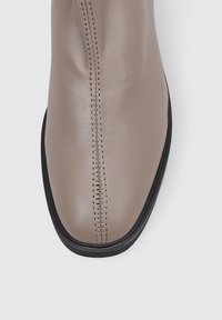 Beige Leder-Ankle-Boot mit runder Zehenpartie, der eine genähte Naht in der Mitte und eine schwarze Gummisohle hat. Glatte Textur.