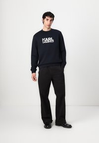 Tamno plava dukserica s bijelim logotipom "KARL LAGERFELD", duži rukavi, rebrasti manžeti, u kombinaciji s crnim hlačama širokih nogavica i crnim cipelama.
