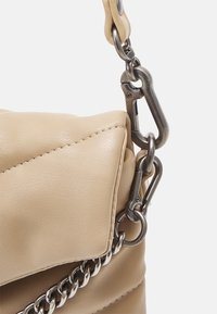 Róg beżowej skórzanej torby z srebrnym łańcuszkiem i metalowymi zapięciami mocującymi pasek do torby na jednolitym tle.