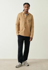 Khaki košile na knoflíky s dvěma předními kapsami a potištěným logem, spárovaná s černými kalhotami a šedými teniskami, na neutrálním pozadí.