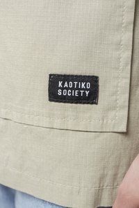 Light beige fabric with a grid texture, featuring a black rectangular label with white text “KAOTIKO SOCIETY” sewn on the edge.