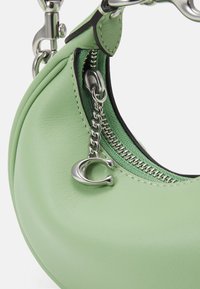 Sac en cuir vert clair en forme de croissant avec une fermeture éclair argentée et une chaînette décorée d'un charme en forme de lettre "C".