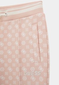 Pantalon de survêtement rose avec une texture douce, comportant un motif de logo répété, une poche latérale et un imprimé « GUESS » sur le devant.