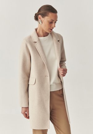 Beige wool coat with a notched lapel, single side pocket, and a relaxed fit. Worn over a light sweater and tan trousers, featuring smooth texture.