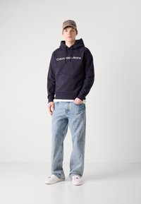Sötét tengerészkék kapucnis pulóver fehér "Calvin Klein Jeans" logóval, világoskék laza fazonú farmernadrággal és fehér sportcipővel párosítva.