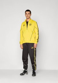 Gul jakke med glidelås og svarte detaljer og logo, kombinert med svarte bukser med gule sidestriper og logo. Sorte treningssko.