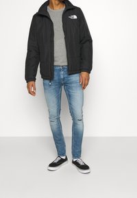 Černá izolovaná bunda s kapucí, šedé tričko s kulatým výstřihem, světle modré dírkované džíny a černé tenisky s bílými podrážkami. Na bundě výrazné logo známé značky.