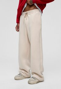 Beige sweatpants gemaakt van een zachte stof, met een wijd-pijpen ontwerp, zijzakken en een koord in de taille. Gepaard met grijze sneakers.